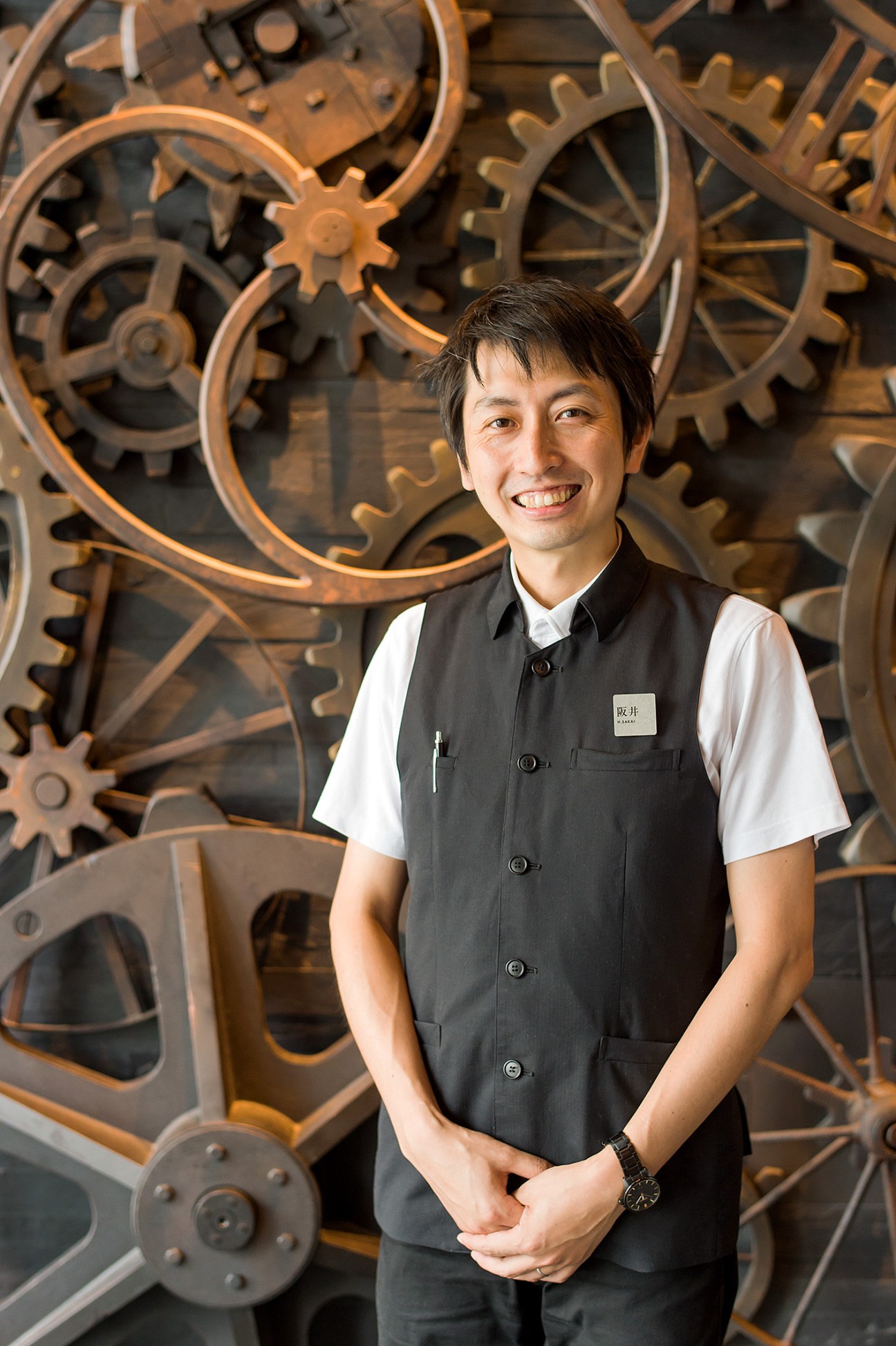 A smiling CCC staff member in uniform stands before a decorative copper gear wall installation.