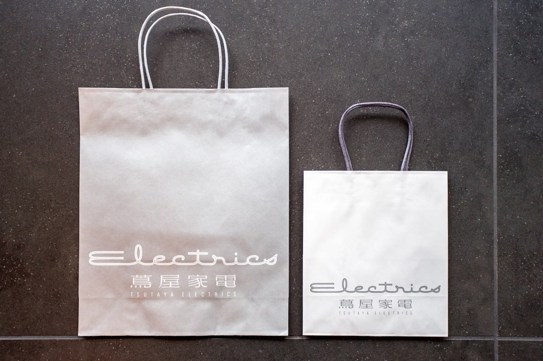 Two white shopping bags with Tsutaya Electrics logo and Japanese characters against a dark textured wall.