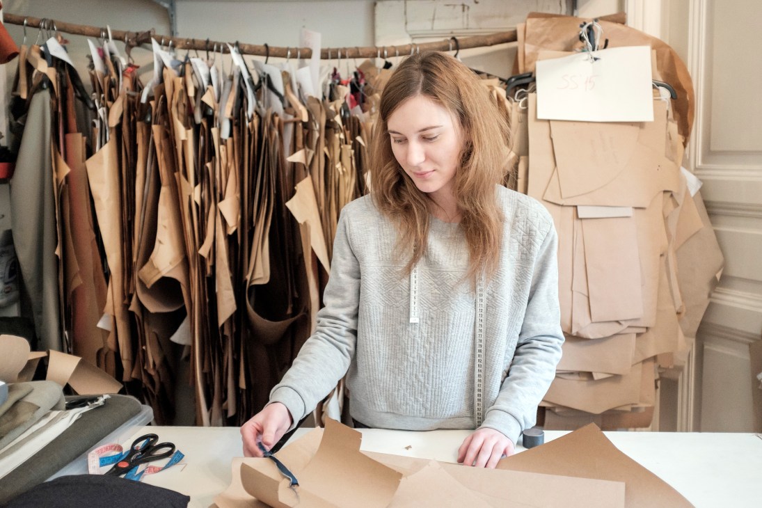 FlatLab assistant Zsuzsanna Tapolcai working at a table in a design studio with fabric samples hanging on a rod behind her.