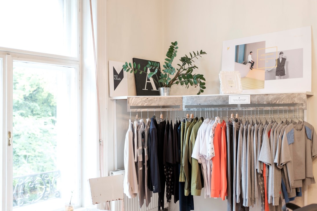 Clothing rack displaying Mei Kawa collection with neutral and colorful garments hung on hangers below a shelf with plants and design materials.