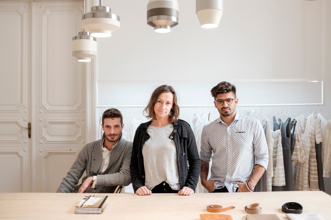 Three members of the Use/unused design team stand at a table in their atelier with clothing racks visible behind them.
