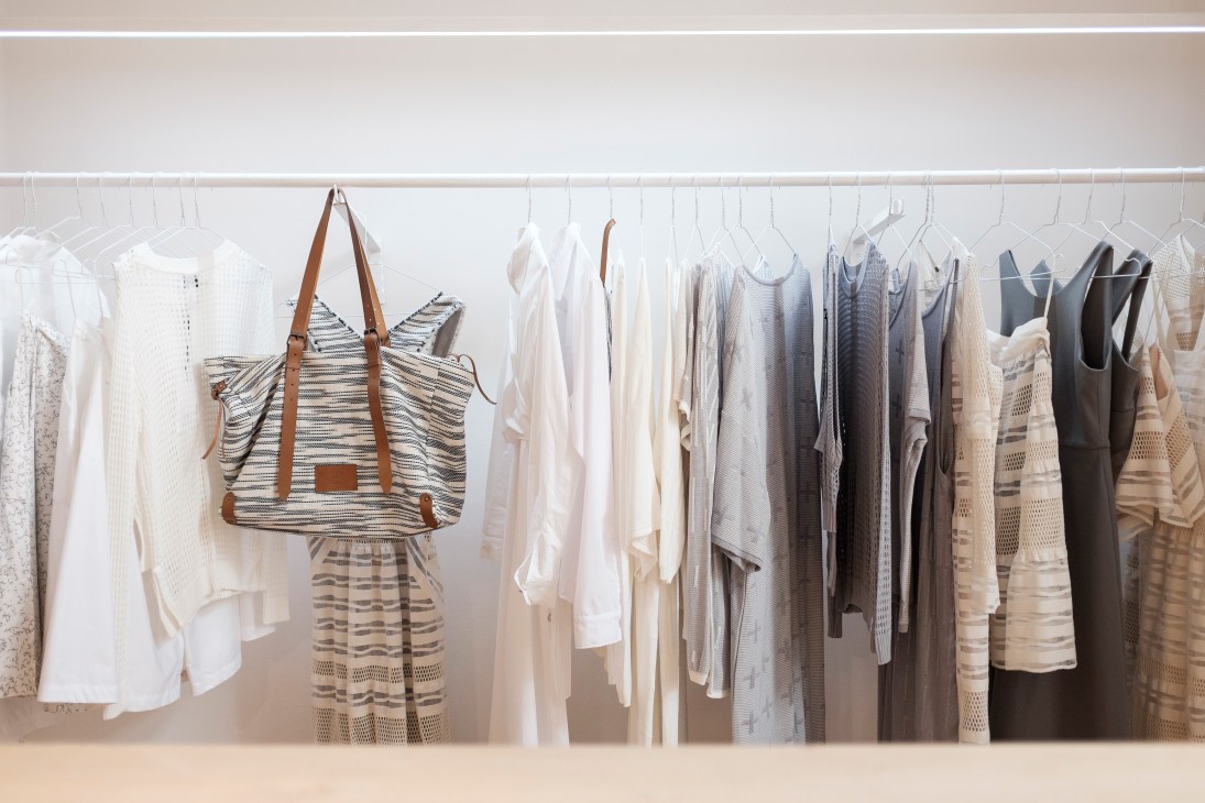 Neutral-toned clothing and a striped tote bag displayed on a white rail in a minimalist showroom.
