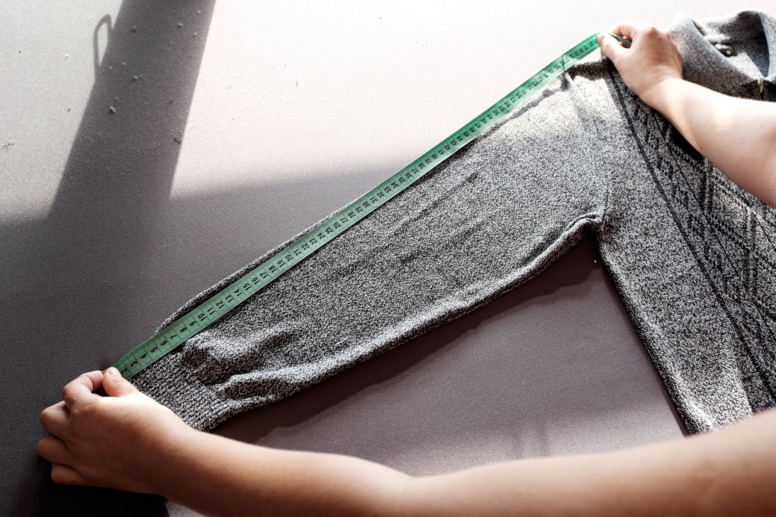 Hands holding a green measuring tape along the edge of grey knitted cardigan fabric.