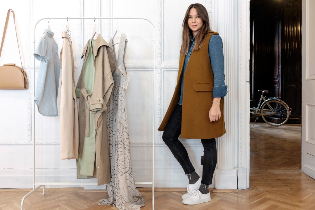 Sandra Sandor, Nanushka founder, standing in a bright showroom with clothing displayed on a rack behind her.
