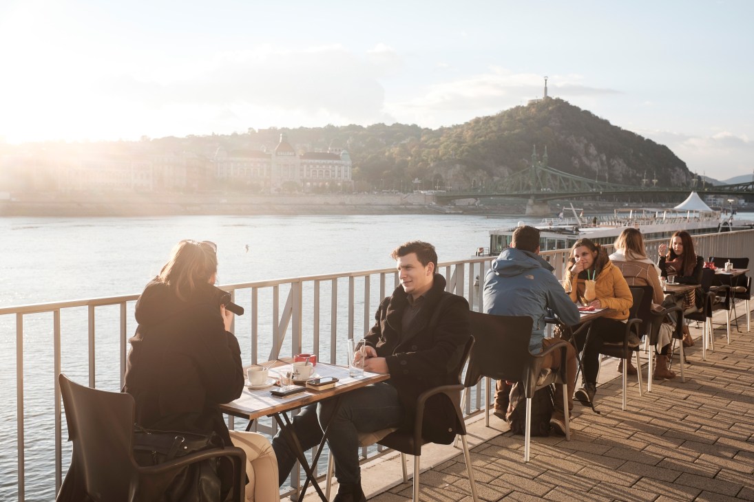 People dining and socializing on a waterfront terrace overlooking the Danube River in Budapest with a bridge and historic buildings visible across the water.