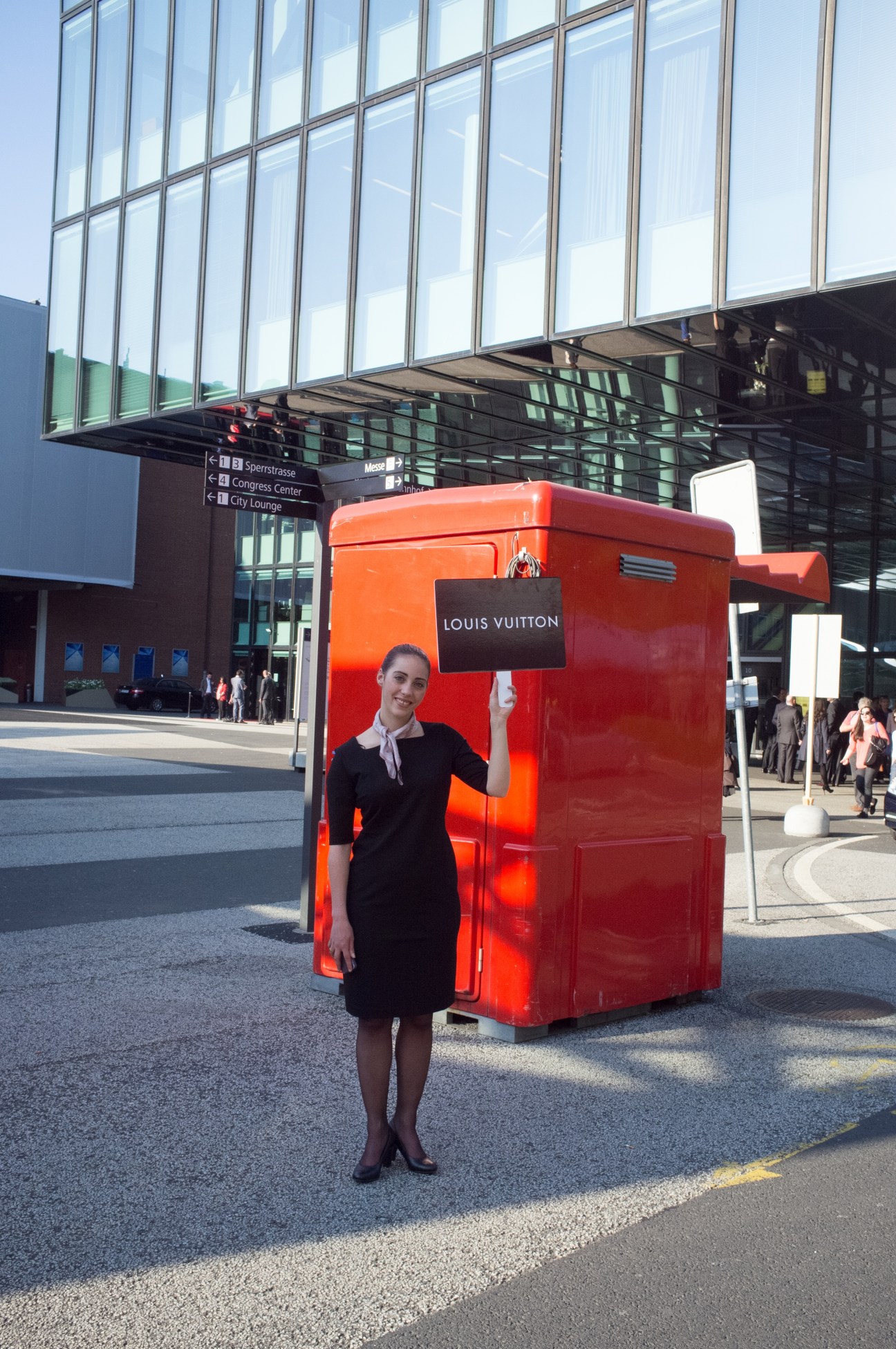 Louis Vuitton staff member in black dress standing beside bright red promotional booth at Baselworld.