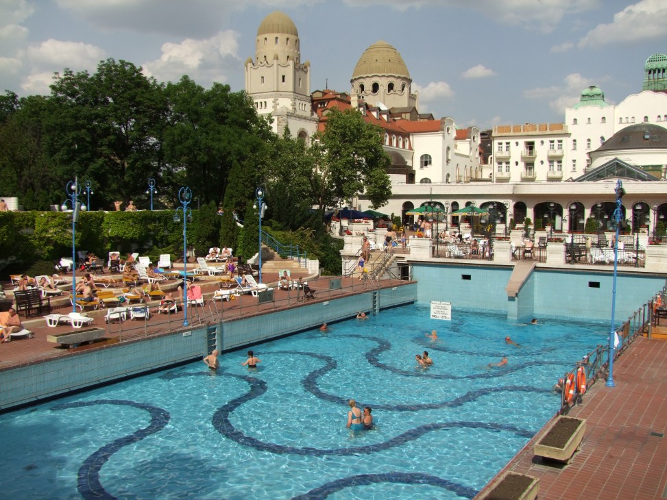 Gellért Baths thermal pool in Budapest with turquoise water, sunbathers on deck, and historic architecture with domed buildings in background.