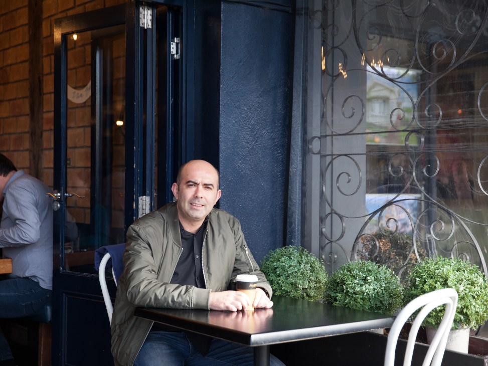 Man in olive jacket sitting at outdoor café table with dark storefront and ornate ironwork behind him.