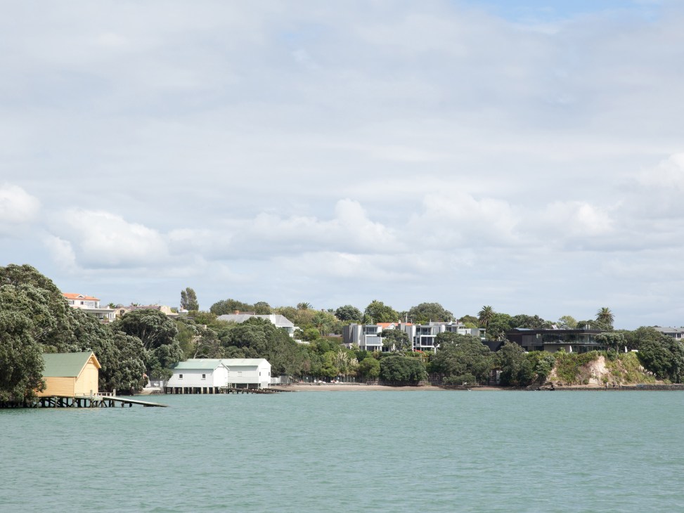 Waterfront boathouses and residential buildings along a turquoise bay with forested hillside
