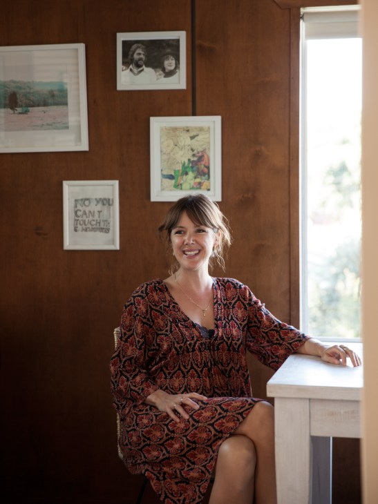 Madeleine Richards smiles while sitting at a table in her home, with framed photographs and artwork displayed on a terracotta-colored wall behind her.
