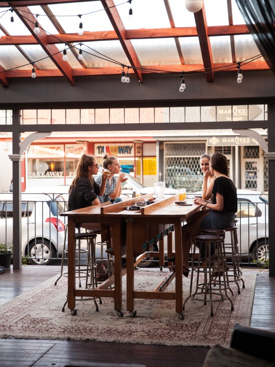 Four women enjoying drinks at a wooden table under a modern pergola with red beams at Tiny Triumphs in Devonport.
