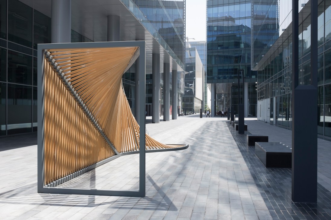 Wooden ribcage-like bicycle rack installation in Dubai Design District's modern glass-fronted courtyard.