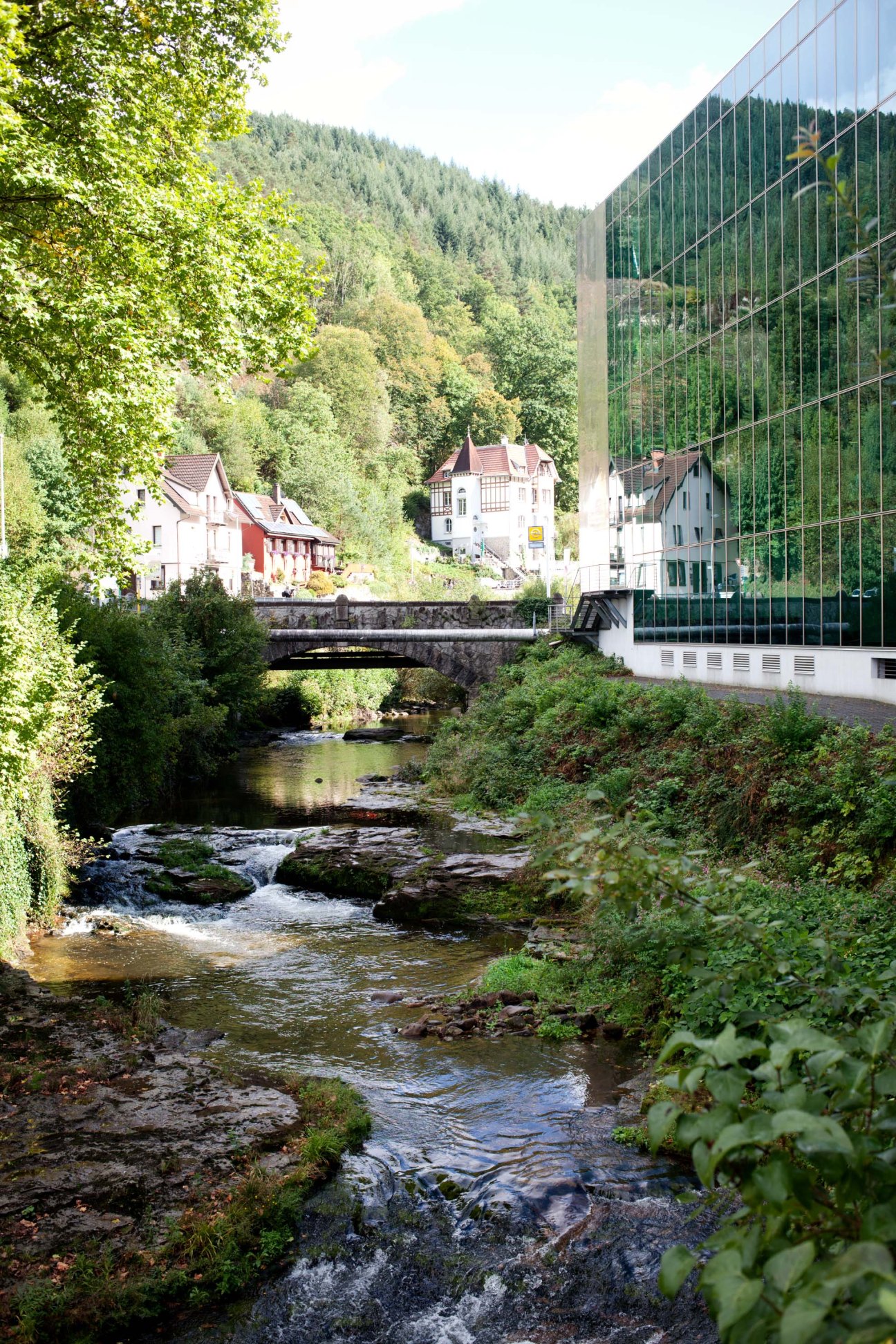A modern glass-walled factory building nestles in a forested Black Forest valley beside a stream and traditional half-timbered houses.