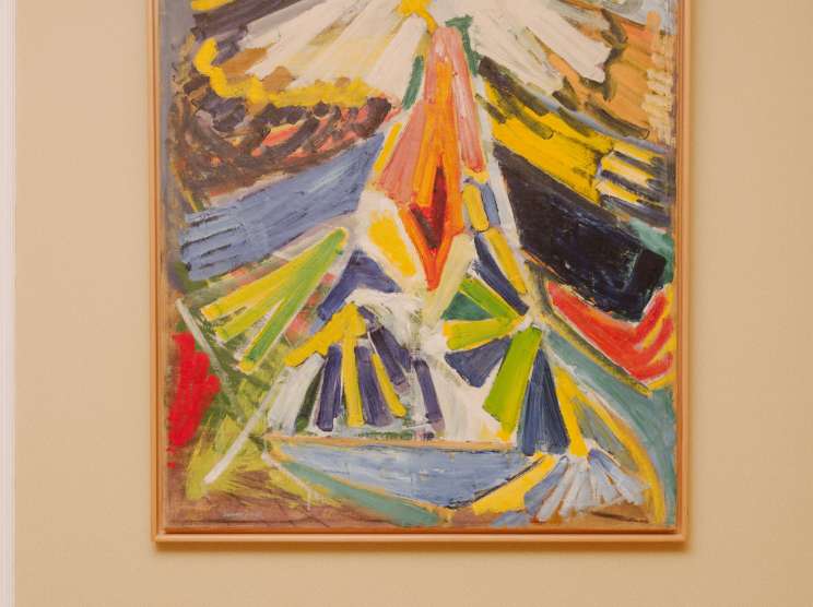 Abstract expressionist painting with bold strokes in yellow, white, red, green, and blue on a cream-colored wall.