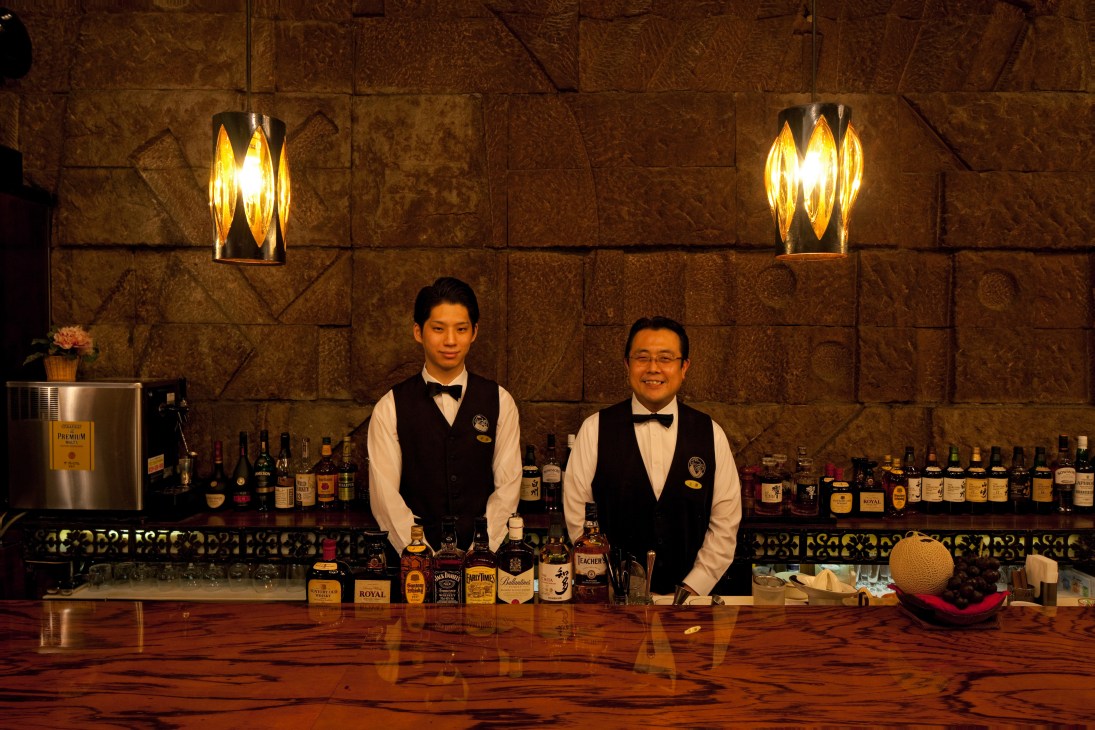 Two bartenders in bow ties and vests stand behind a wooden bar lined with whisky bottles.