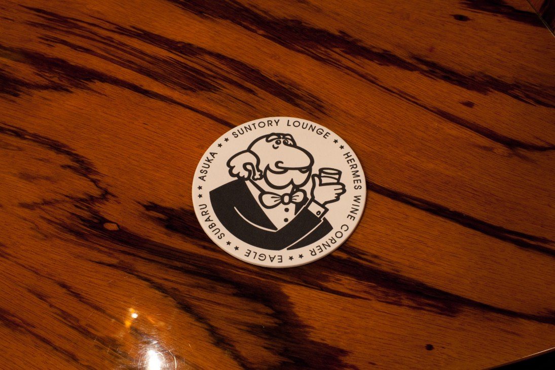 Circular coaster for Suntory Lounge Eagle featuring an illustrated character holding a drink on wood surface.