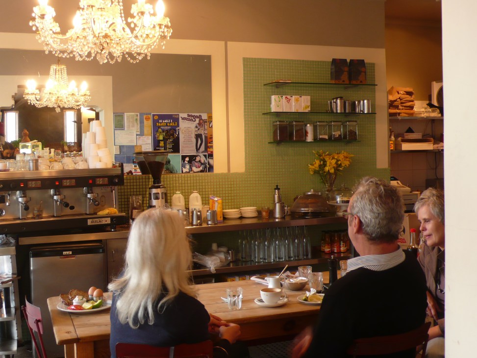 Interior of The Earth Food Store cafe in Bondi with customers seated at wooden table, green tiled wall, shelving, and chandelier lighting.