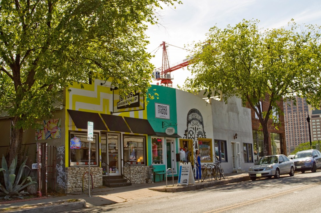 Colorful storefronts with yellow and green painted facades line a tree-shaded street in East Austin.