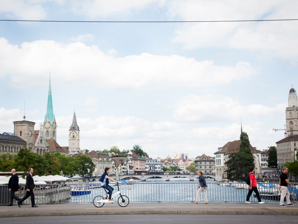 Cyclist riding a white Gocycle G2 electric bike along Zurich's riverfront with historic churches and buildings in background.