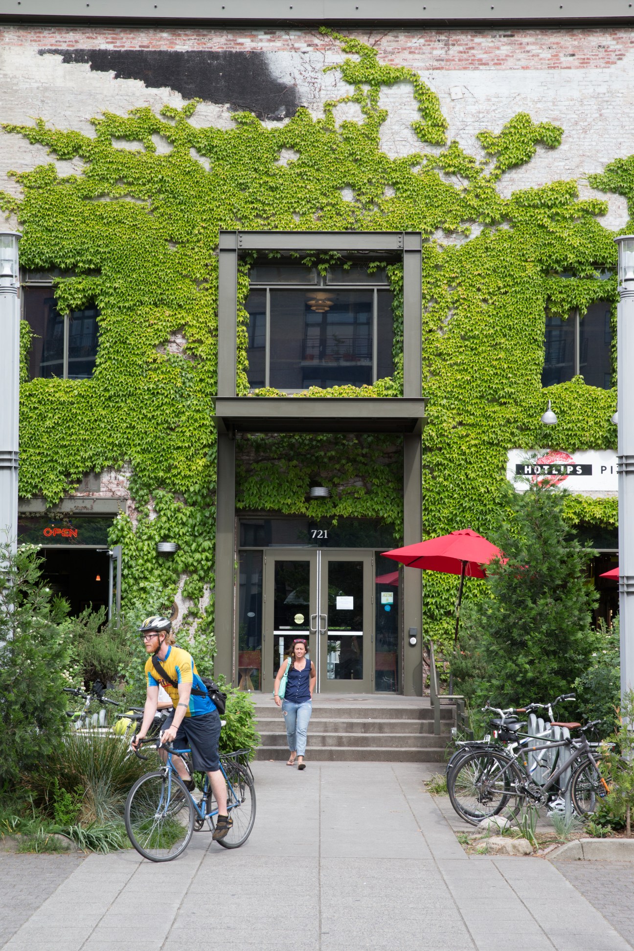 Ivy-covered entrance to a modern building with green umbrellas and cyclists passing by on the street.