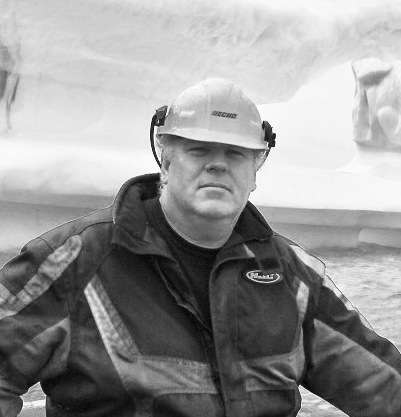Ed Kean, a sea captain wearing a hard hat and dark jacket, stands on a boat in black and white.