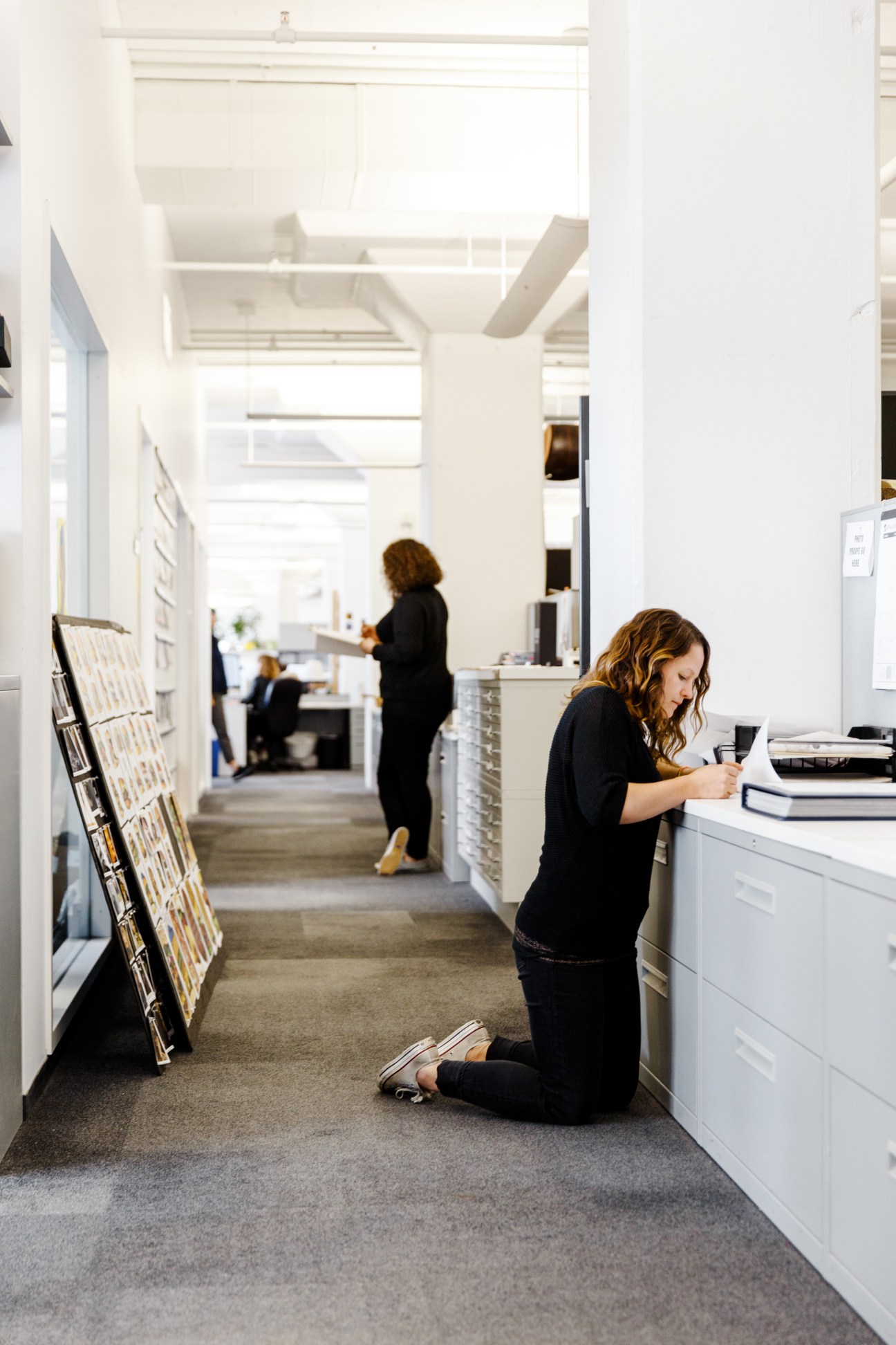 Photo editor Gabrielle Plucknette kneeling in a white hallway at New York magazine's office, reviewing materials.