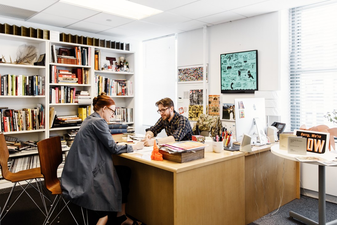 Two people work at a desk in a bright office with floor-to-ceiling bookshelves and artwork on the walls.