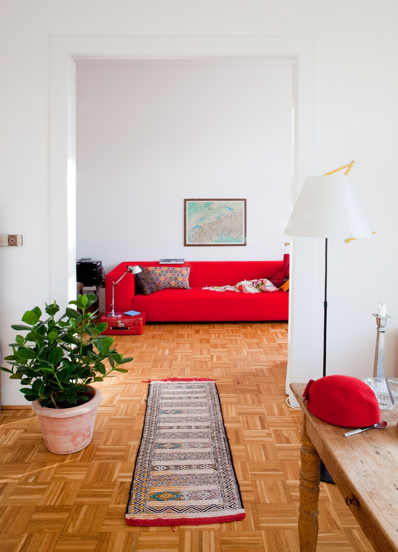 Bright apartment interior with red sofa, wooden herringbone flooring, potted plant and geometric rug.