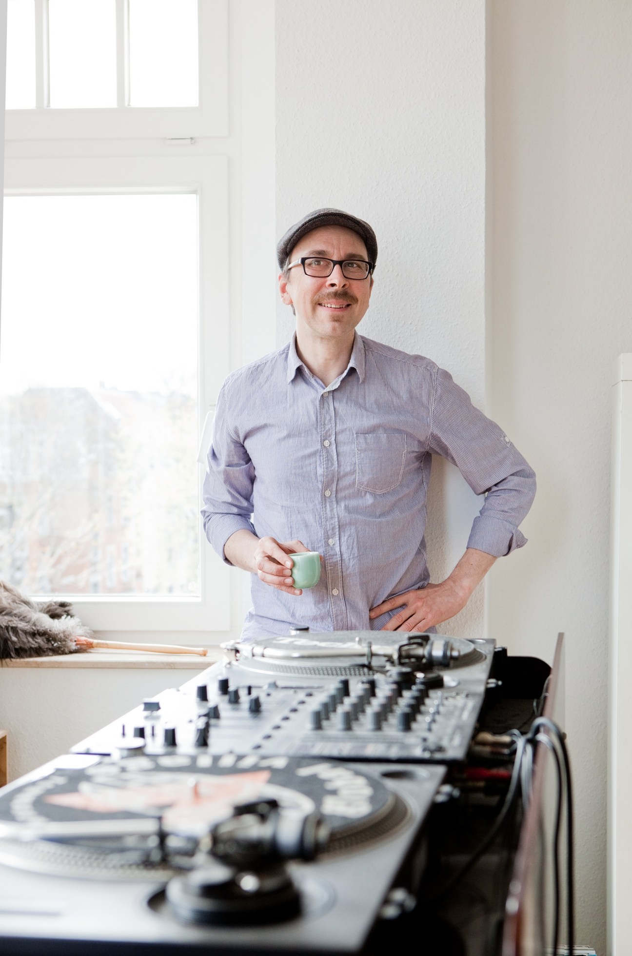 Hans Nieswandt, a DJ and writer, stands in a bright room holding a cup, with DJ equipment on a table in front of him.