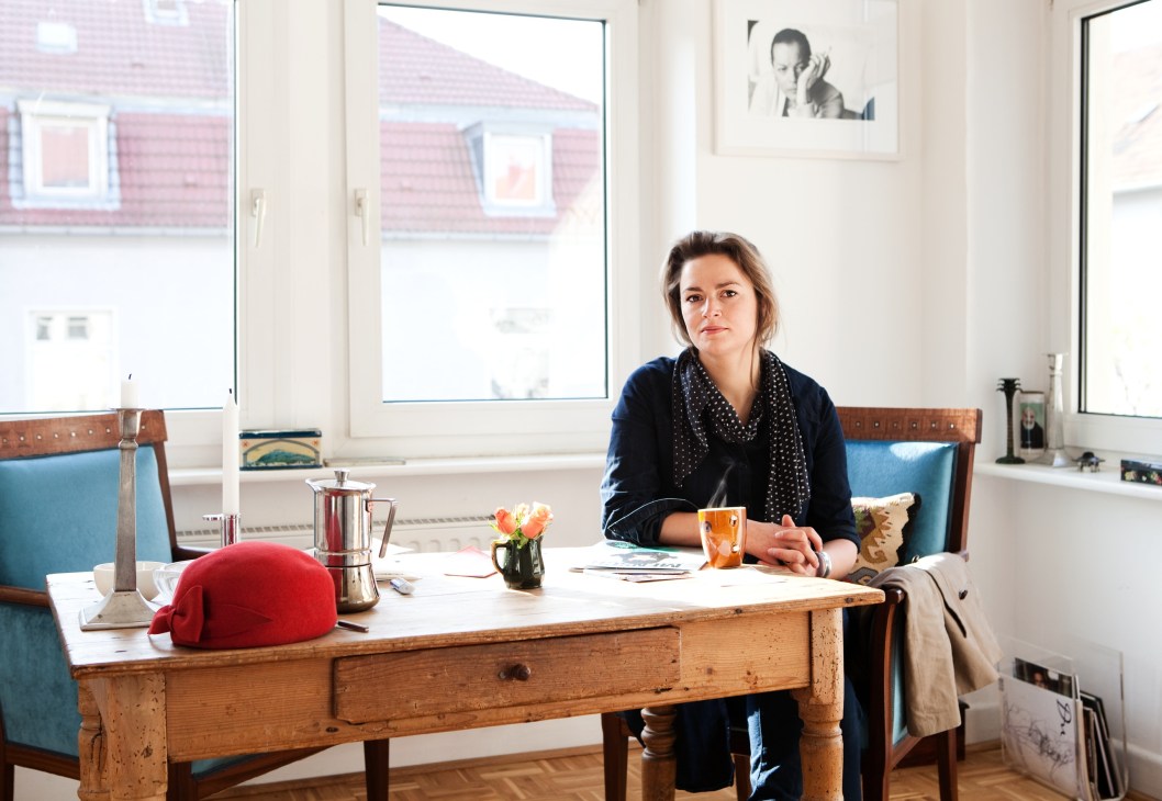 Designer Julia Wülfing sitting at a wooden desk in her bright apartment with blue-upholstered chairs and large windows overlooking red-roofed buildings.