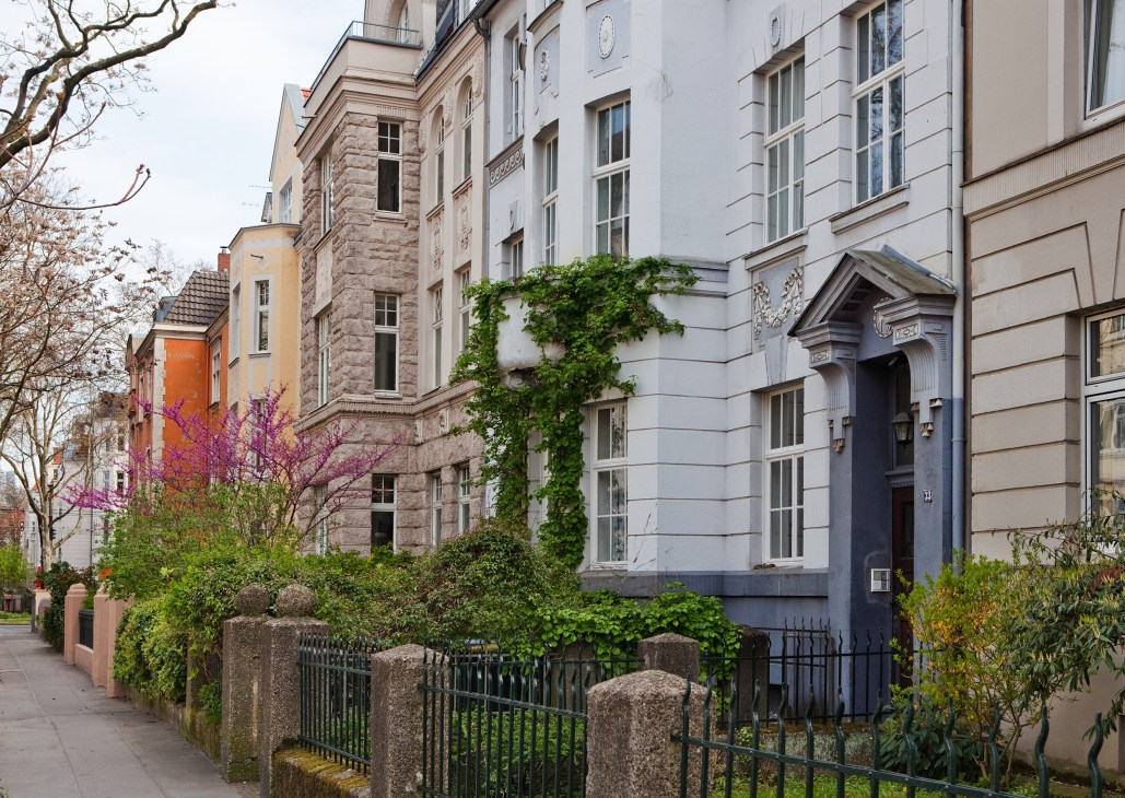 Tree-lined residential street with historic early 1900s apartment buildings featuring ivy-covered facades and ornamental architecture.
