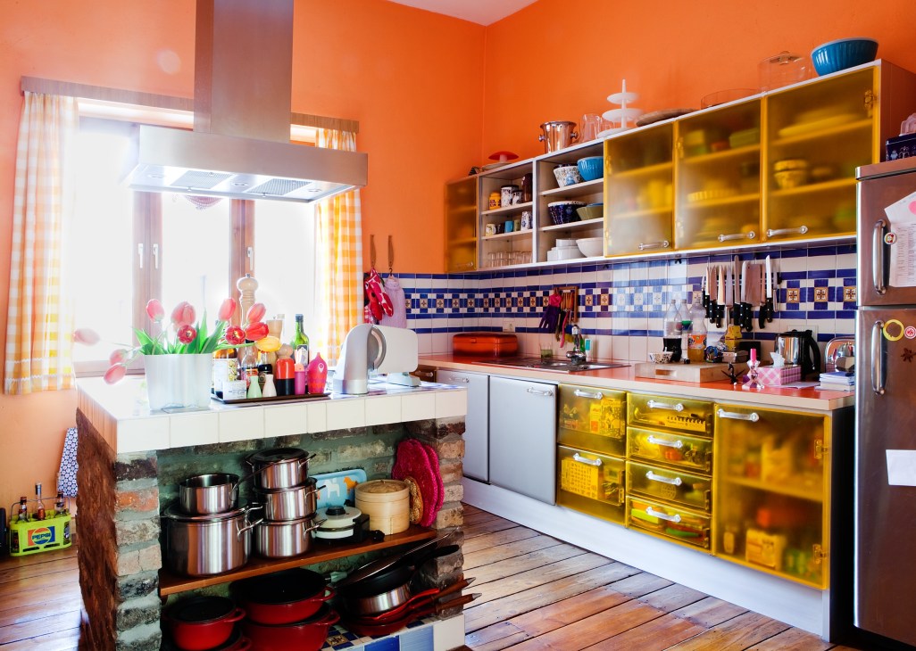 Bright orange kitchen with yellow cabinets, blue and white tile backsplash, and exposed brick base in Achim Schmitz's home.