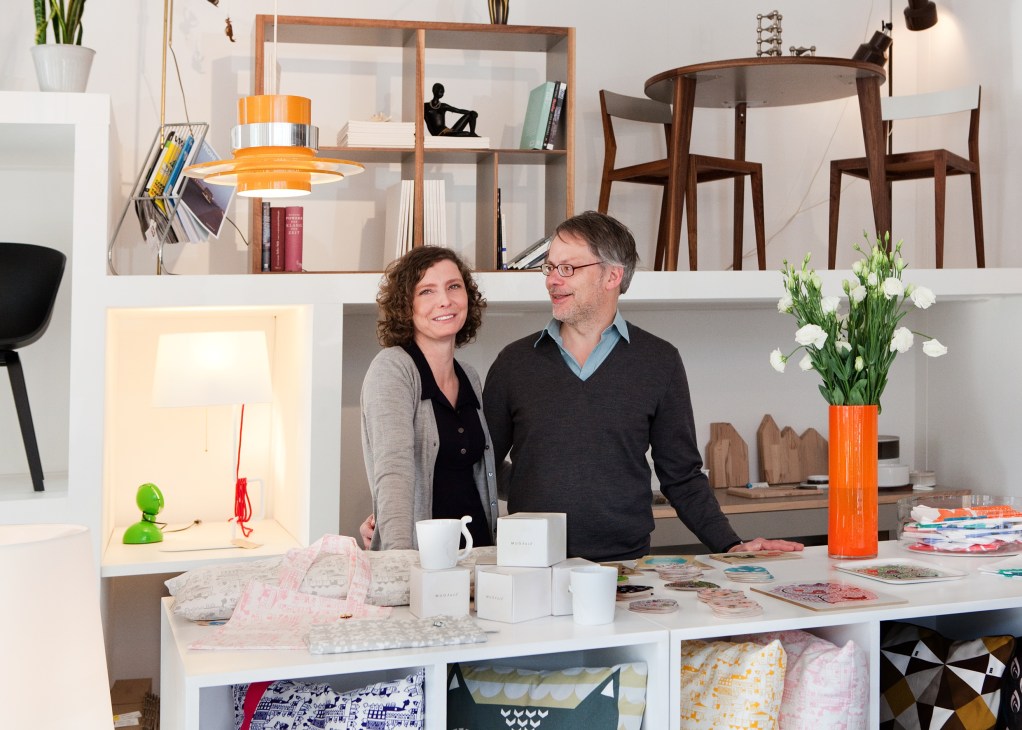 A man and woman stand at a white counter in a modern interior design shop displaying home goods and décor items.