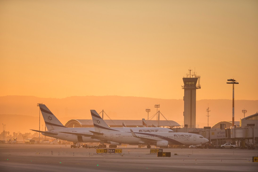 El Al aircraft parked at Ben Gurion airport terminal during sunrise with control tower and hazy sky.