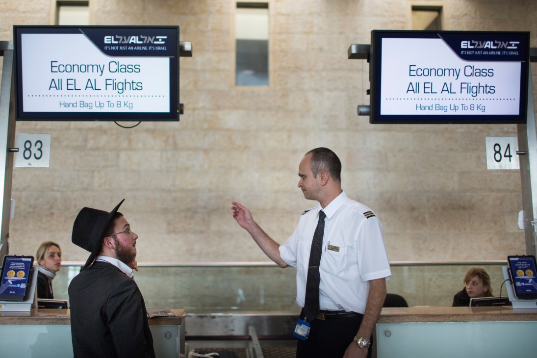 El Al airline staff member assists a passenger at Ben Gurion airport check-in counter with overhead signage displaying economy class flight information.