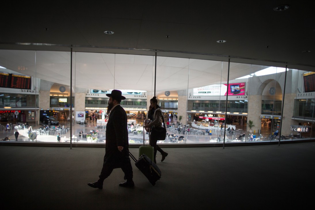 Travellers with luggage walk through Ben Gurion airport terminal past large windows overlooking the main hall.