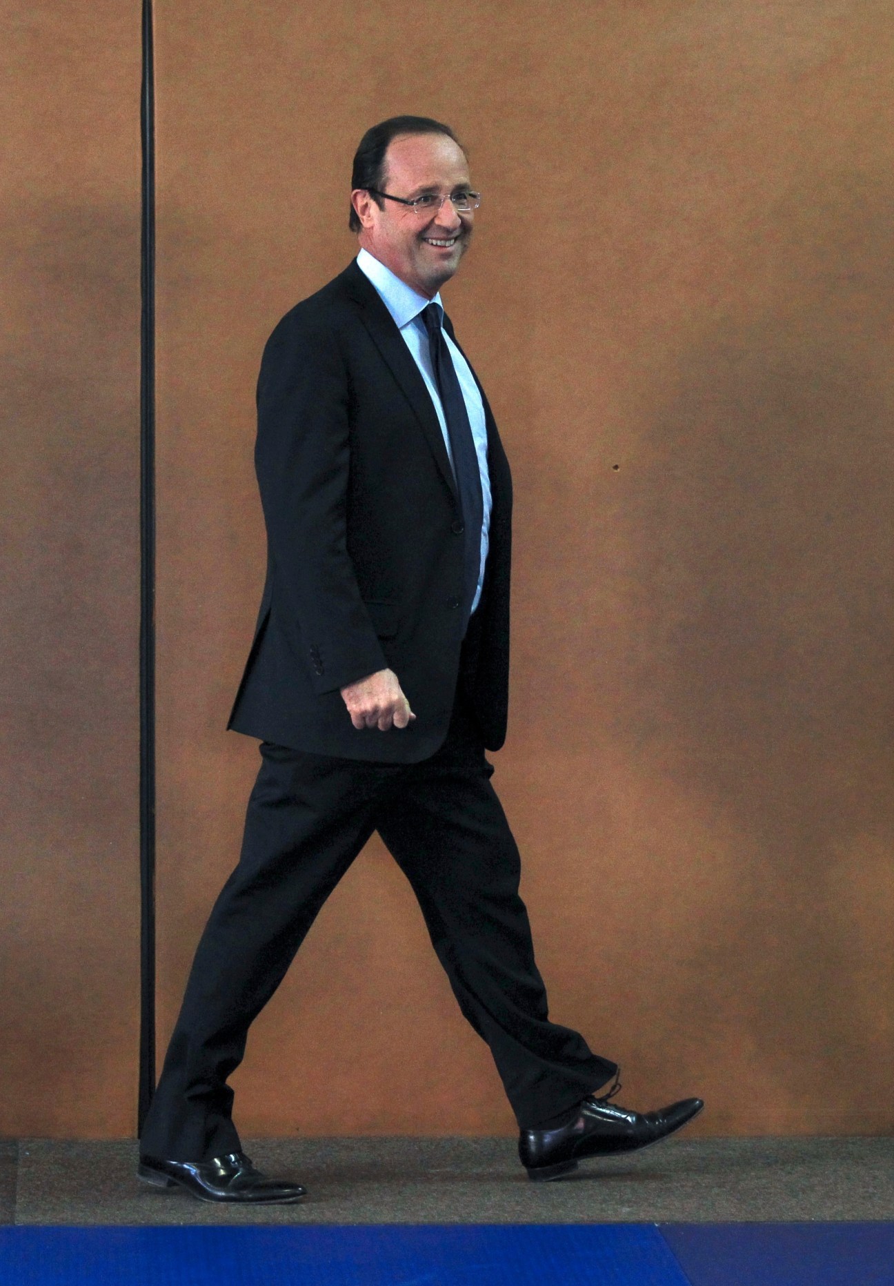 François Hollande walking in a dark suit and light blue shirt against a beige wall.