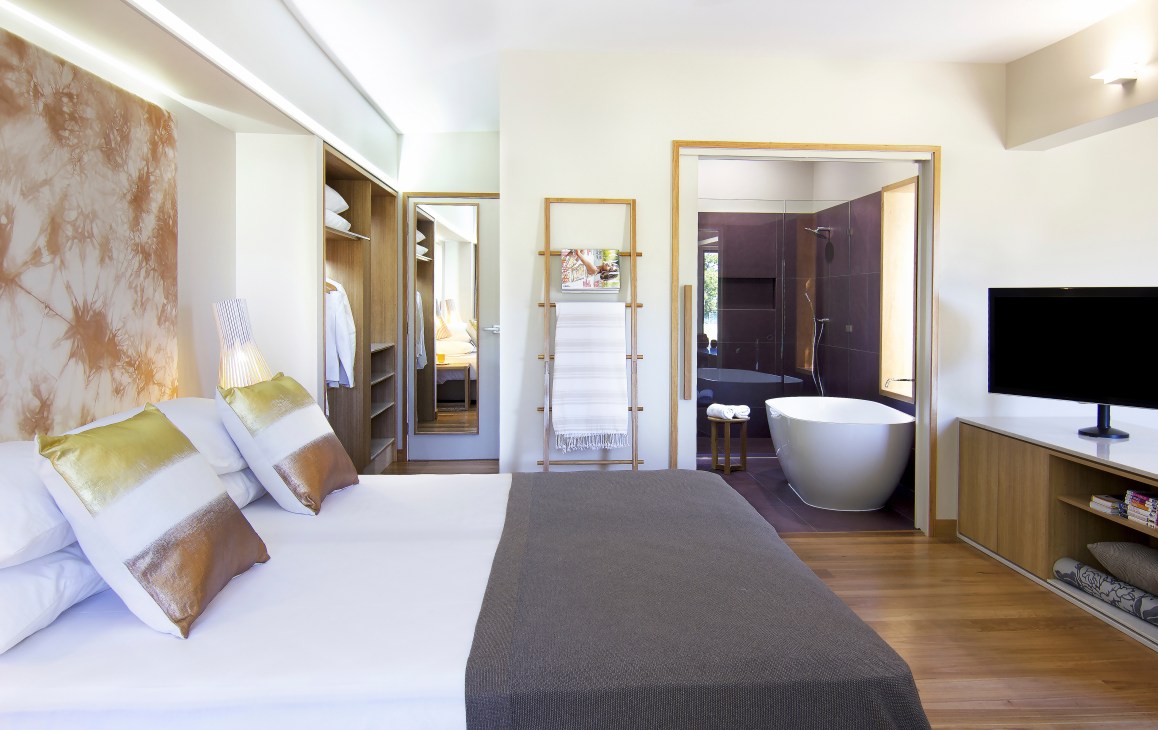 Modern hotel bedroom with white bed, gold pillows, open ensuite bathroom with freestanding tub, and wood accents.