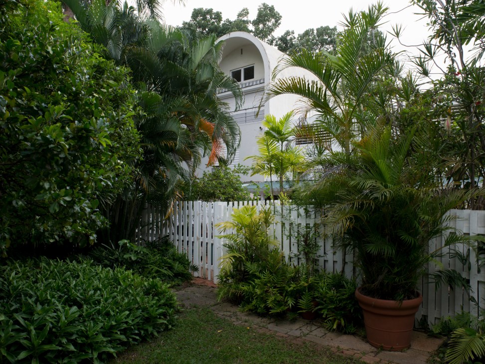 White modernist townhouse with curved architectural details surrounded by lush tropical vegetation and white picket fence.