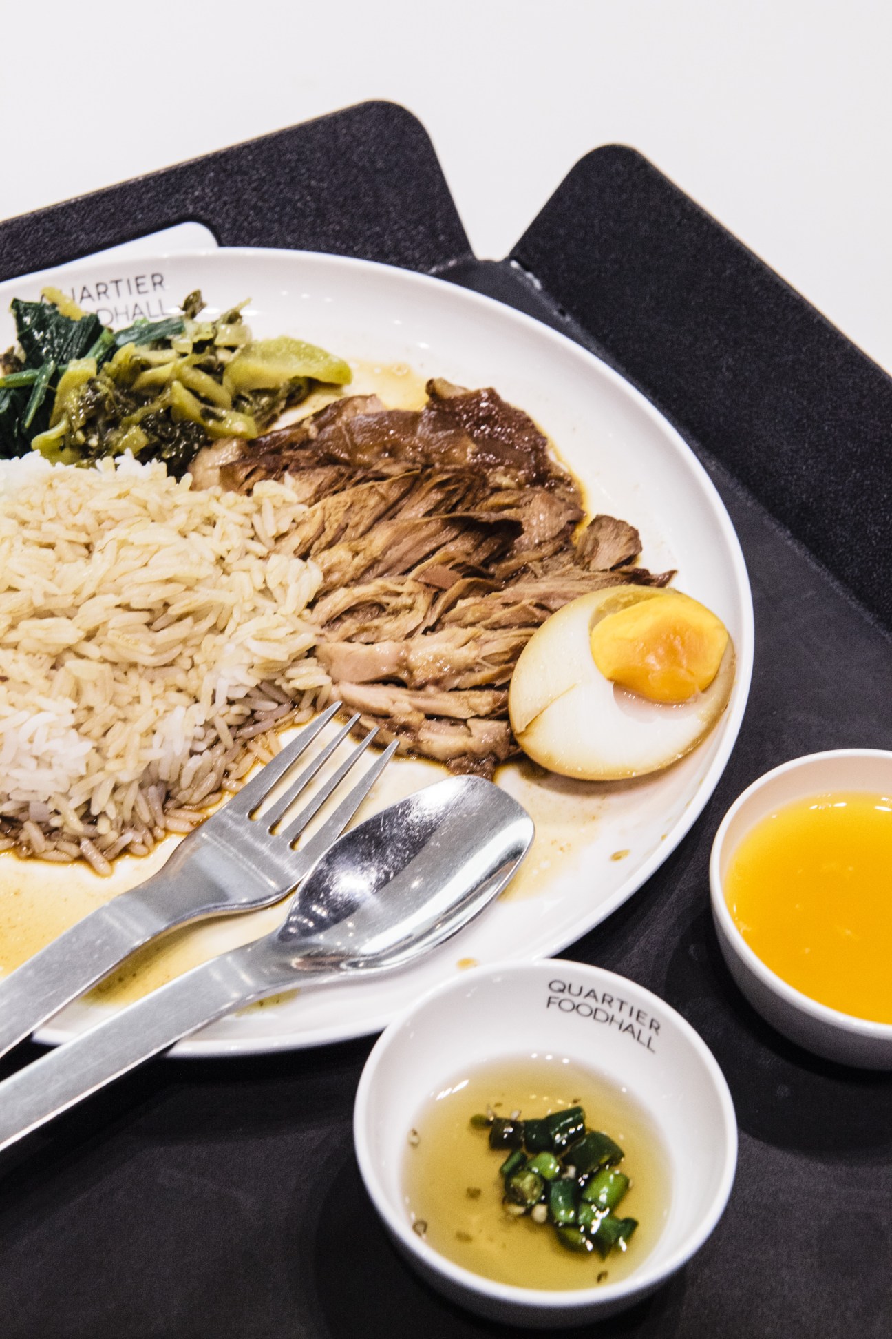 Plated dish at Quartier Food Hall featuring sliced meat, rice, egg and greens with sauce and condiments.