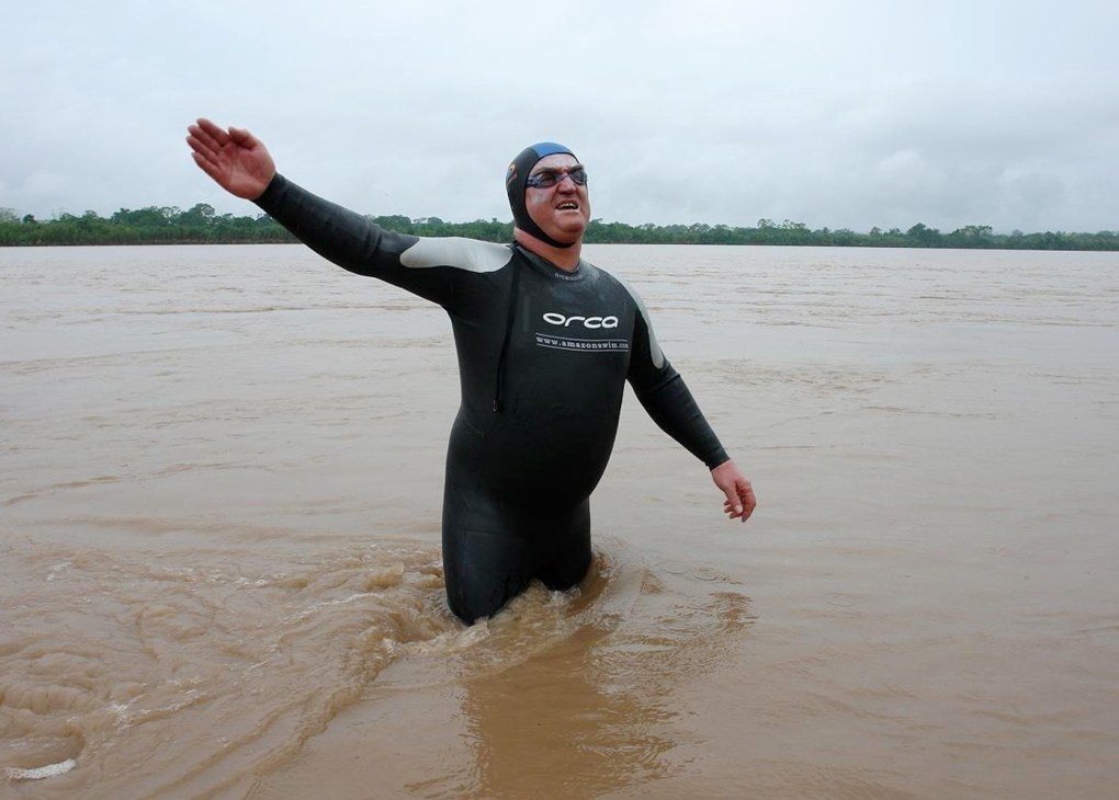 Martin Strel wading in the Amazon River in Peru, wearing a black wetsuit and waving, with rainforest visible in the background.