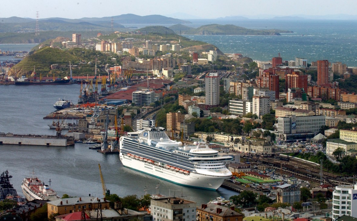 Aerial view of Vladivostok's port city with a large cruise ship docked in the harbor, surrounded by buildings and mountains.