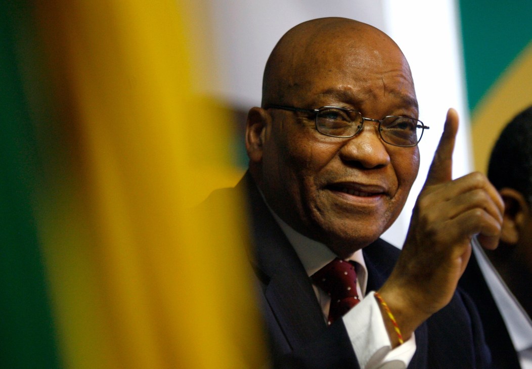 Jacob Zuma gestures while speaking, with South African flag colors blurred in the background.