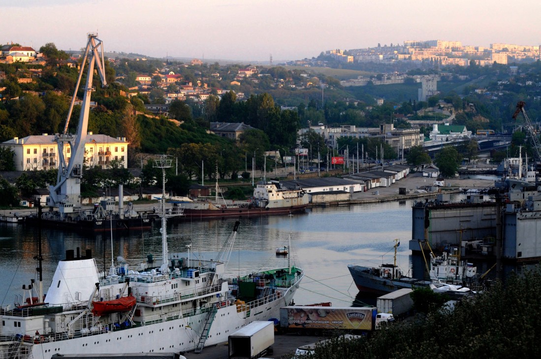 Industrial port with ships docked, cranes, and hillside cityscape in background.
