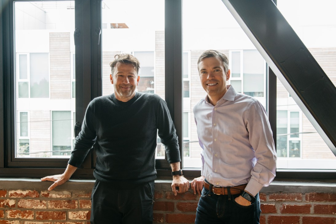Two men standing by large windows in an office space with exposed brick, one in dark sweater, one in light shirt