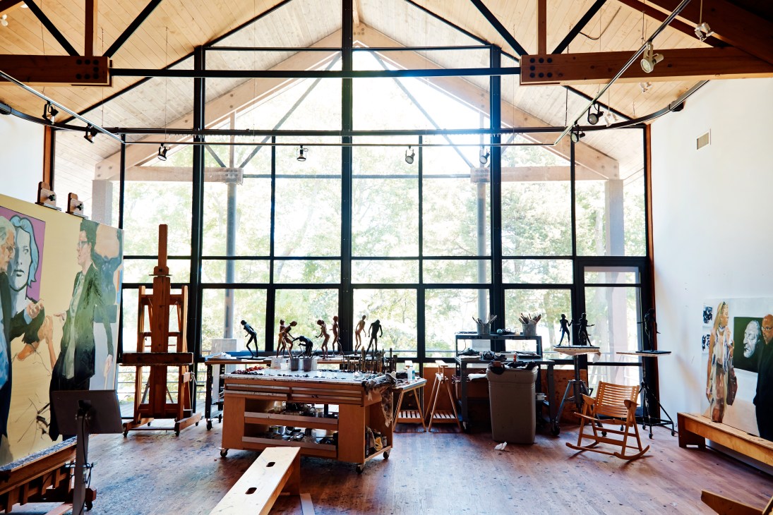 A spacious artist's studio with high ceilings, large windows, wooden work tables, and paintings on the walls.