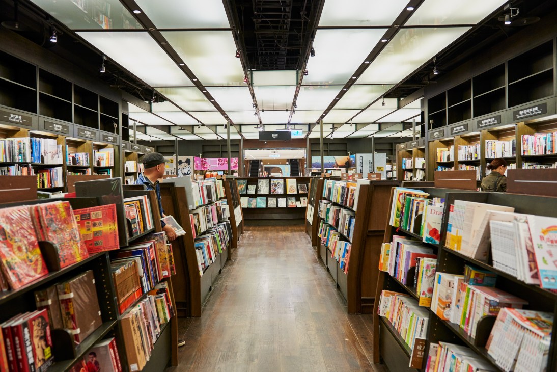 Interior of Eslite bookstore showing wooden shelving units and customers browsing books under bright ceiling lights.