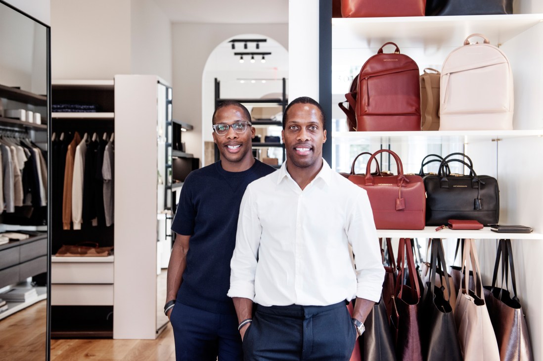 Two men pose inside a minimalist retail store with leather bags and accessories displayed on white shelves behind them.