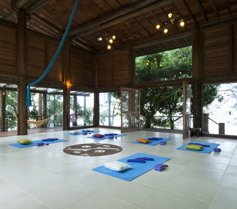 Spacious yoga studio with blue mats, colorful cushions, wooden beams, and ocean views through large windows.