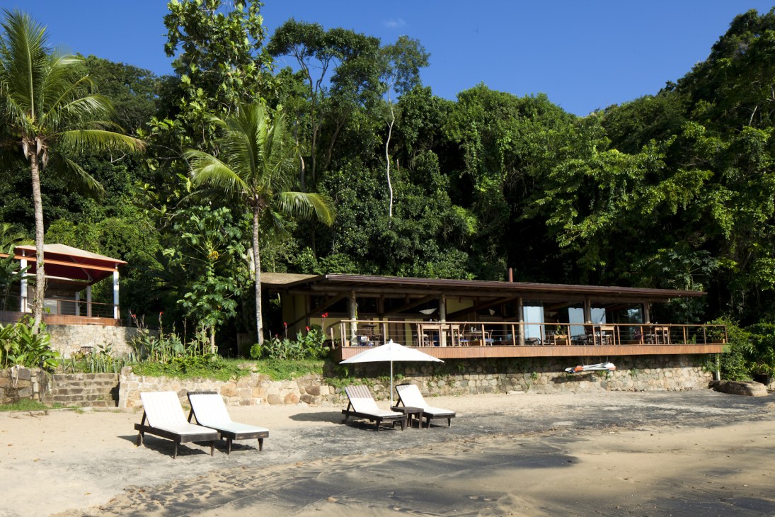 Beach resort with wooden lodge nestled against lush jungle, lounge chairs on sand at The Island Experience, Ilha Grande, Brazil.